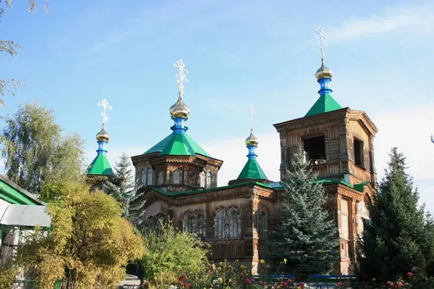 Eine große hölzerne orthodoxe Kirche mit grünen Dächern und goldenen Zwiebeltürmen, umgeben von Bäumen und Blumen unter blauem Himmel.