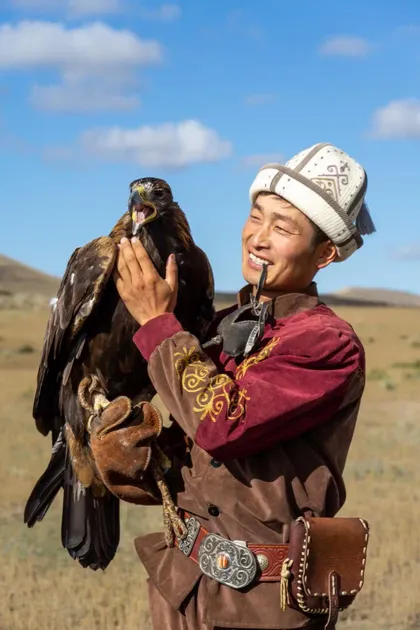 Ein lächelnder Mann in traditioneller Kleidung hält einen großen goldenen Adler auf seiner behandschuhten Hand auf einem Feld.