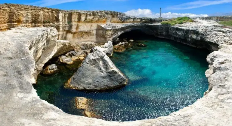 Una grotta circolare piena di acqua turchese limpida, circondata da scogliere rocciose di colore chiaro sotto un cielo azzurro.