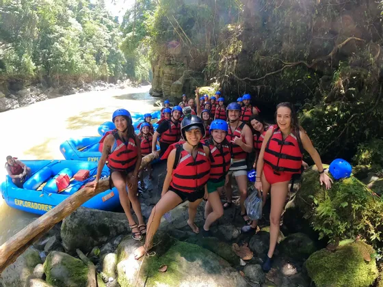 Un gruppo WeRoad in viaggio, che indossa giubbotti di salvataggio rossi e caschi blu, posa per una foto su una sponda rocciosa del fiume accanto ai loro gommoni.