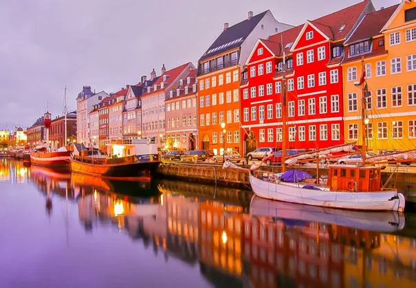 Edificios coloridos y barcos amarrados reflejados en un tranquilo canal de la ciudad bajo un cielo crepuscular.