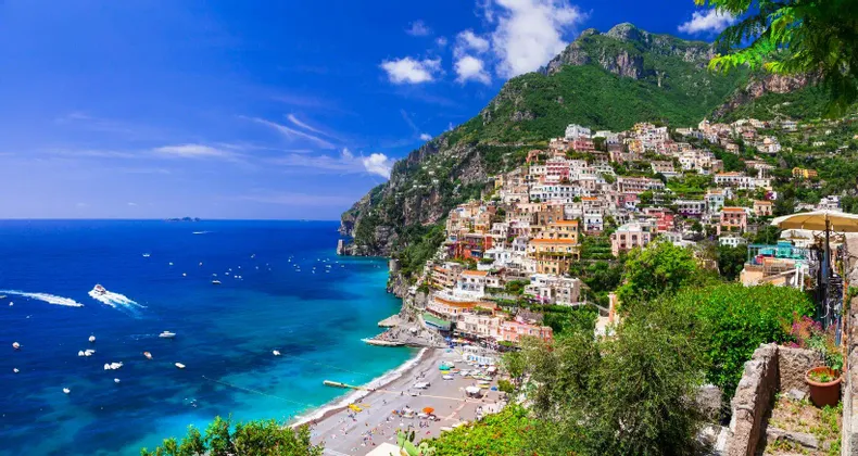 Ein farbenfrohes Dorf, das in eine grüne Klippe gebaut wurde und einen Strand sowie ein türkisfarbenes Meer mit Booten unter blauem Himmel überblickt.