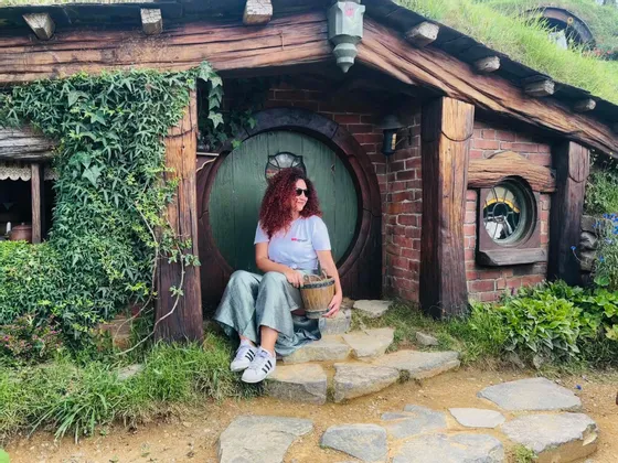 Una donna con i capelli rossi ricci siede sui gradini di pietra di una casa in collina con una porta verde rotonda, tenendo un secchio di legno.