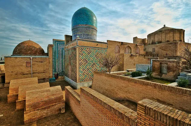Un complesso di storici edifici in mattoni con una prominente cupola a piastrelle blu e intricati mosaici geometrici sotto un cielo nuvoloso.