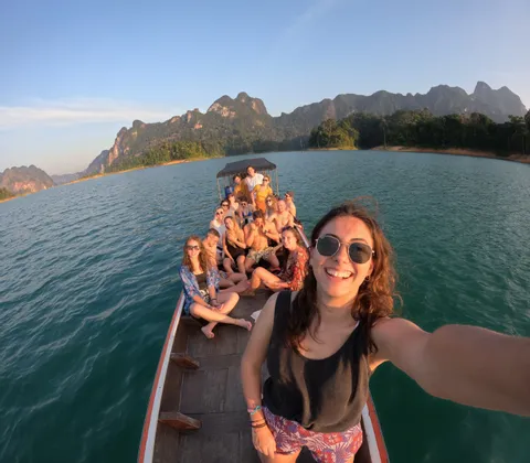 Una mujer se hace un selfie con un viaje en grupo WeRoad en una barca de cola larga en un lago rodeado de montañas.