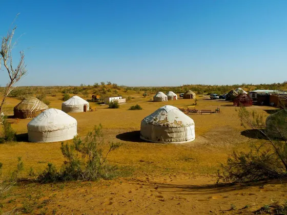 Un accampamento di yurte tradizionali sparse in un paesaggio sabbioso e arido con bassi arbusti verdi sotto un cielo azzurro e limpido.