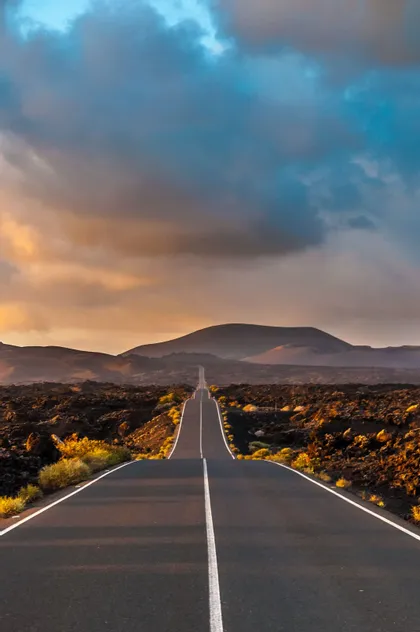 Una strada diritta si estende attraverso un paesaggio vulcanico roccioso verso le montagne sotto un cielo nuvoloso al tramonto.