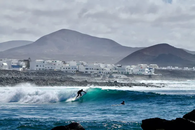 Un surfista cavalca una grande onda di fronte a una località costiera con edifici bianchi e colline vulcaniche sotto un cielo nuvoloso.