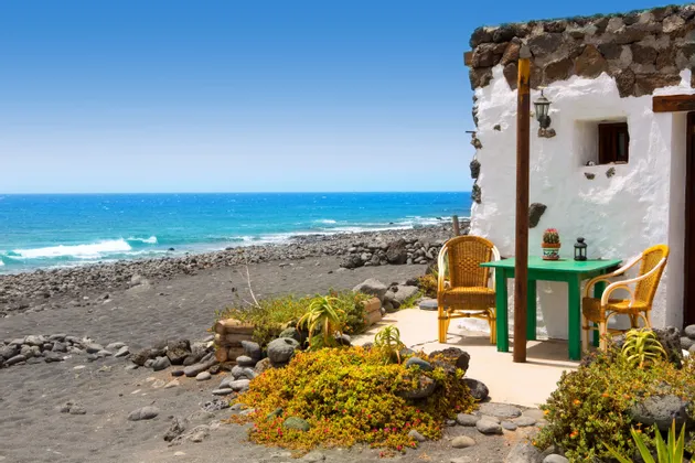 Un patio con un tavolo verde e due sedie di vimini all'esterno di una casa imbiancata su una spiaggia di sabbia nera accanto all'oceano.
