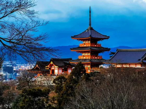 Una pagoda e un tempio giapponese arancioni a più livelli, su una collina che domina una città, incorniciati da alberi spogli sotto un cielo azzurro.