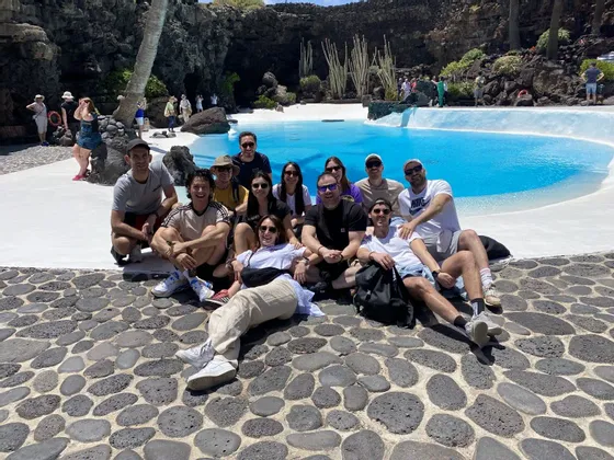 Un gruppo WeRoad è riunito su un patio di ciottoli scuri per una foto di gruppo, con una piscina azzurra brillante e rocce vulcaniche sullo sfondo.
