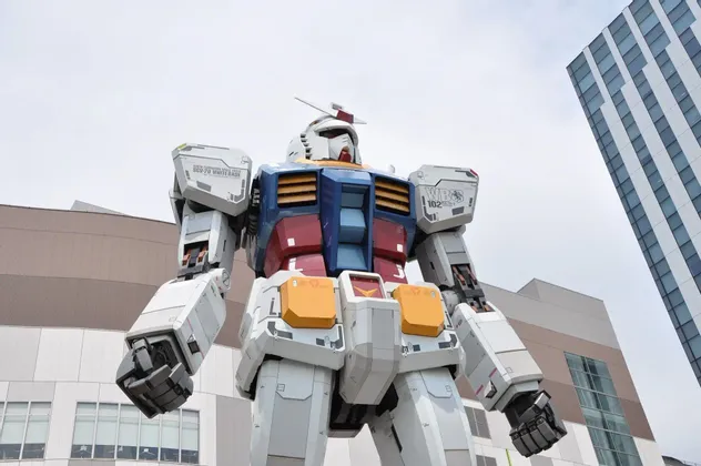 Una inquadratura dal basso di una statua gigante a grandezza naturale del robot Gundam, posta di fronte ad alti edifici moderni.