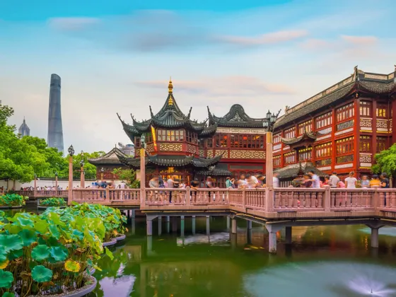 Una pagoda tradizionale cinese su uno stagno con foglie di loto, con un moderno grattacielo sullo sfondo e persone su un ponte.