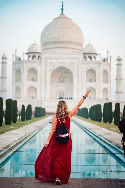 Eine Frau von hinten in einem langen roten Kleid und einem WeRoad-Rucksack hebt ihren Arm vor dem Taj Mahal und seinem Spiegelbecken.