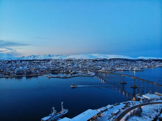 Vista aérea de una ciudad costera nevada al anochecer, con un puente largo sobre el agua y montañas al fondo.