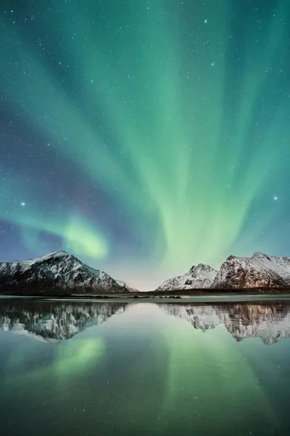 Grüne Polarlichter leuchten über schneebedeckten Bergen, die sich in einem ruhigen Gewässer spiegeln.