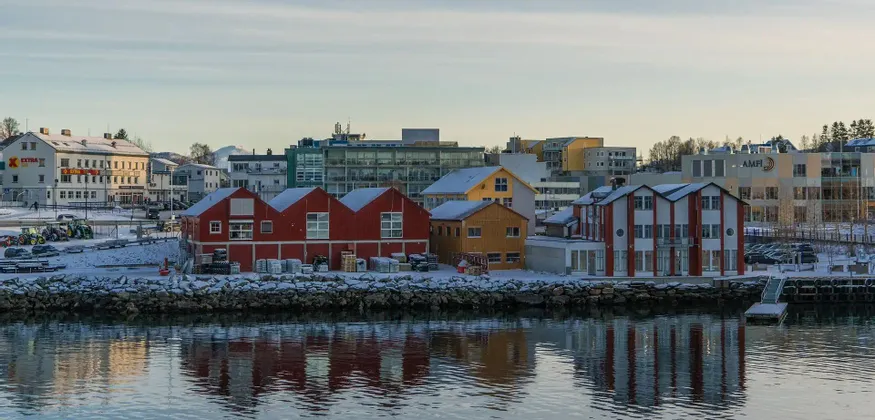 Edificios coloridos con tejados nevados se alinean junto a un paseo marítimo, sus reflejos visibles en el agua tranquila.