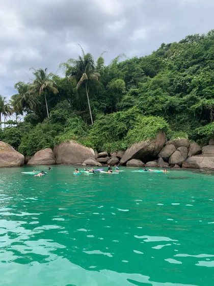 Un viaggio di gruppo WeRoad galleggia su tavole da surf in acque calme e turchesi vicino a una costa rocciosa e lussureggiante sotto un cielo nuvoloso.