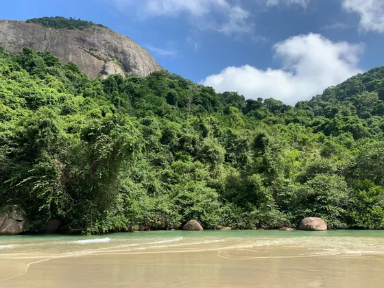Une plage de sable avec des eaux calmes et vert clair au pied d'une montagne luxuriante couverte de jungle sous un ciel bleu.