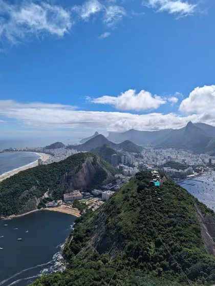 Vue plongeante d'une ville côtière avec des plages et des montagnes verdoyantes bordant l'océan sous un ciel bleu et nuageux.