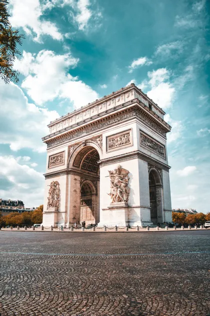 Un grande e ornato arco trionfale si erge su un'ampia piazza lastricata sotto un cielo azzurro con nuvole sparse.