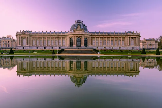 Un grande edificio in stile classico con una cupola centrale perfettamente riflessa in una pozza d'acqua immobile sotto un cielo dai toni violacei.