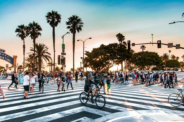 Un trafficato attraversamento pedonale con pedoni e ciclisti su una strada fiancheggiata da alte palme, sotto un cielo al tramonto.