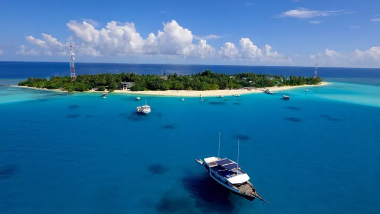 Luftaufnahme einer tropischen Insel mit einem weißen Sandstrand, umgeben von türkisfarbenem Wasser und mehreren vor Anker liegenden Booten.