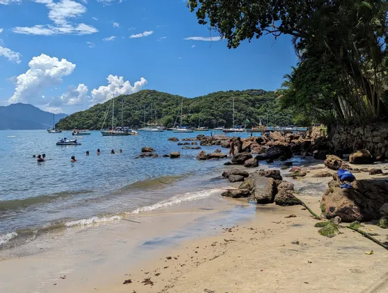 Une vue depuis une plage de sable et de rochers, avec des gens qui nagent dans une baie où des voiliers sont ancrés, et une colline verdoyante en arrière-plan.