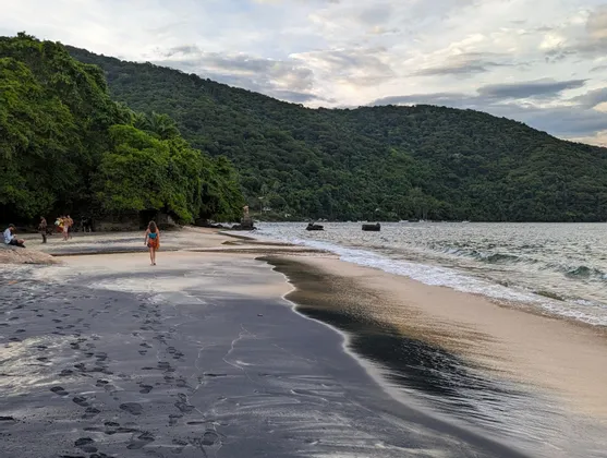 Une femme marche sur une plage de sable sombre au pied d'une grande colline boisée sous un ciel nuageux.