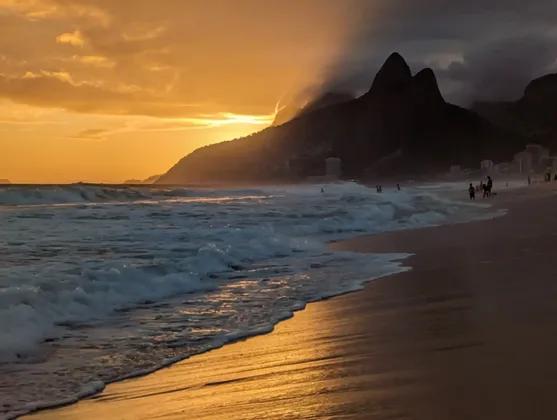 Les vagues de l'océan s'écrasent sur une plage de sable au coucher du soleil, avec une lumière dorée se reflétant sur l'eau et des montagnes en silhouette à l'arrière-plan.