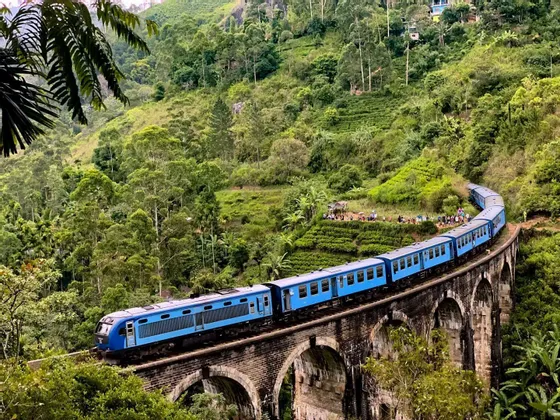 Un tren azul viaja a través de un gran puente de piedra arqueado que serpentea por un exuberante y verde valle tropical con laderas aterrazadas.