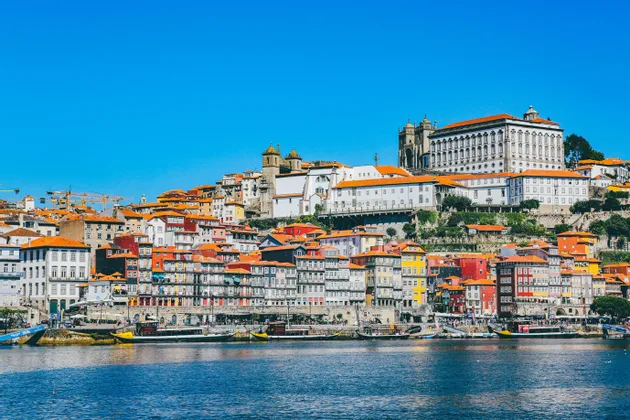 Un paesaggio urbano di edifici colorati con tetti di tegole rosse su una collina che domina un fiume sotto un cielo azzurro chiaro