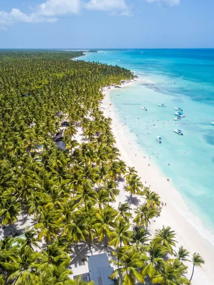 Una toma aérea muestra una larga playa de arena blanca bordeada de palmeras, con personas y barcos en el océano turquesa claro.