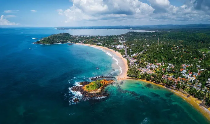 Vista aerea di una costa tropicale con una spiaggia a mezzaluna, un isolotto roccioso e acqua turchese sotto un cielo parzialmente nuvoloso.