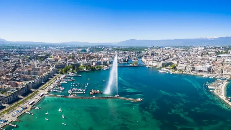 Vue aérienne d'une grande fontaine jaillissant de l'eau d'une baie turquoise, avec un paysage urbain dense et des montagnes lointaines.