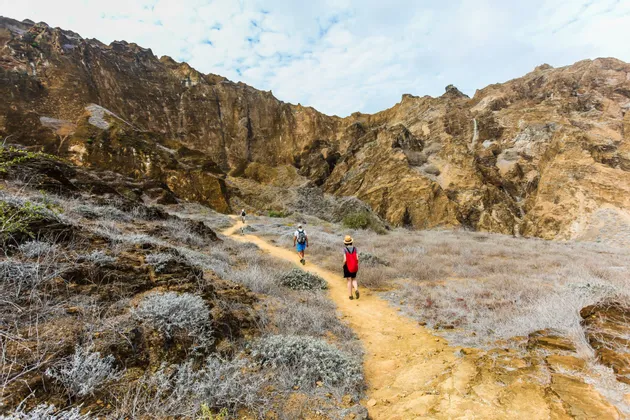 Eine WeRoad-Gruppenreise wandert auf einem schmalen Feldweg durch ein felsiges Tal, mit steilen Klippen im Hintergrund.