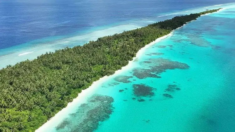 An aerial view of a long, narrow tropical island covered in palm trees, with a white sand beach, turquoise reef, and deep blue ocean.