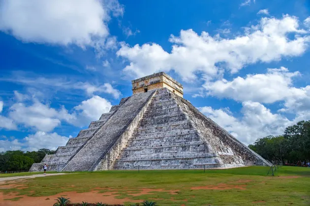 Una grande piramide a gradoni di pietra si erge su un campo erboso sotto un cielo azzurro brillante con nuvole bianche sparse.