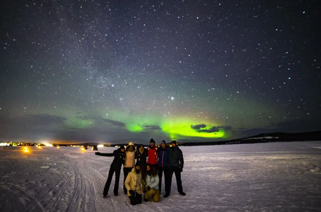 Un gruppo WeRoad in viaggio posa su una piana innevata di notte, con l'aurora boreale e le stelle visibili nel cielo.