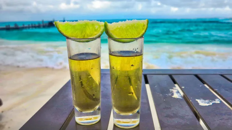 Due shot di tequila con spicchi di lime salati poggiano su un tavolo con vista su un oceano turchese e una spiaggia sabbiosa.