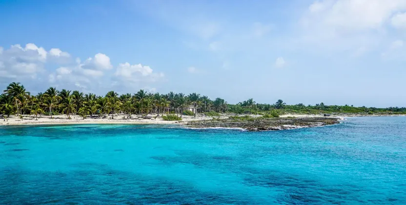Una costa tropicale con palme e spiaggia sabbiosa, bagnata da acque limpide e turchesi sotto un cielo parzialmente nuvoloso.