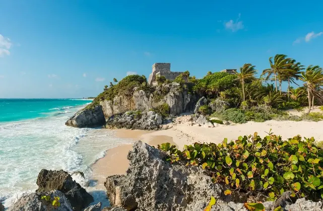 Un'antica rovina in pietra sorge su una scogliera verde a picco su una spiaggia sabbiosa, con un'iguana che riposa su una roccia vicino al mare turchese.