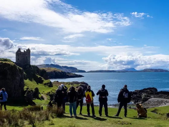 Un gruppo WeRoad su una costa erbosa, ammirando le rovine di un castello su una scogliera a picco sul mare.