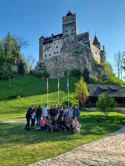 Un gruppo WeRoad in viaggio posa per una foto in una giornata di sole davanti a un castello storico, arroccato su una verde collina rocciosa.