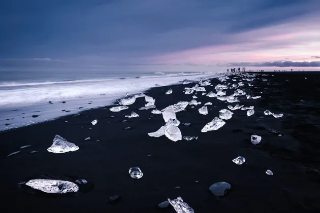 Un viaggio di gruppo WeRoad su una spiaggia di sabbia nera dove blocchi di ghiaccio sono sparsi dalle onde dell'oceano sotto un cielo crepuscolare.