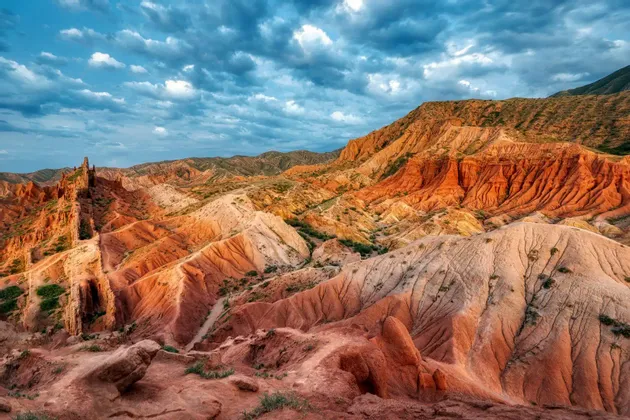 Una vista amplia de un paisaje árido de formaciones de cañones rojos y naranjas bajo un cielo azul nublado.
