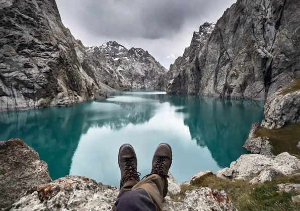 Una vista en primera persona de botas de senderismo en una cornisa rocosa, con vistas a un lago tranquilo de color turquesa rodeado de montañas empinadas bajo un cielo nublado.