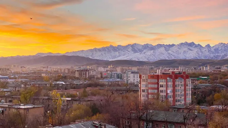 Un paisaje urbano con edificios anidados frente a una cordillera de montañas nevadas bajo un cielo de atardecer dorado.