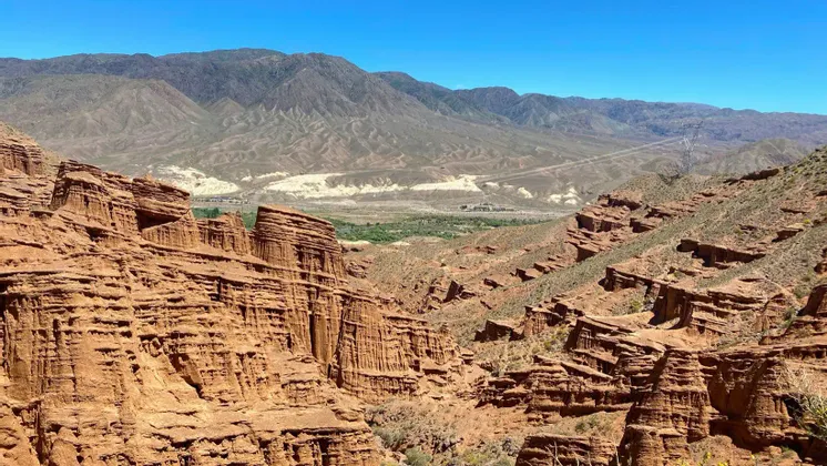 Una vista a un cañón con formaciones rocosas rojas en capas, con montañas secas distantes bajo un cielo azul brillante.